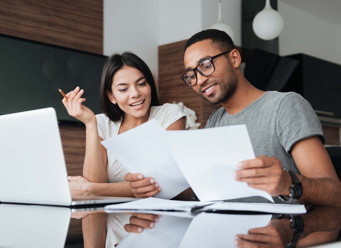 Annuities - Cheerful Young Couple Using a Laptop Together at the Kitchen Table While Analyzing Financial Documents